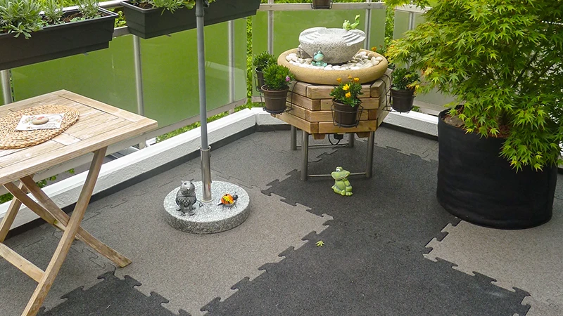 Na fotografii je útulný balkon pokrytý dvoubarevnými puzzle dlaždicemi WARCO. Malá fontána, dřevěná police a několik květináčů vytvářejí příjemnou atmosféru. Podlaha poskytuje vysoký komfort díky elastickému, protiskluzovému povrchu a je zároveň odolná vůči povětrnostním vlivům. Díky přesnému systému spojování lze přidat barevné akcenty, které prostoru dodají moderní vzhled.