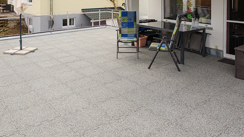 Na střešní terase jsou položeny šedě mramorované terasové desky WARCO z PU vázaného gumového granulátu. Jejich puzzle spoj zaručuje přesné napojení a kombinuje odolnost vůči počasí s dlouhou životností. Stabilní materiál tlumí kročejový hluk a přispívá k čistému, upravenému dojmu. Navíc poskytuje protiskluzový povrch a vysoký komfort při chůzi, což dělá tuto podlahu velmi praktickou.