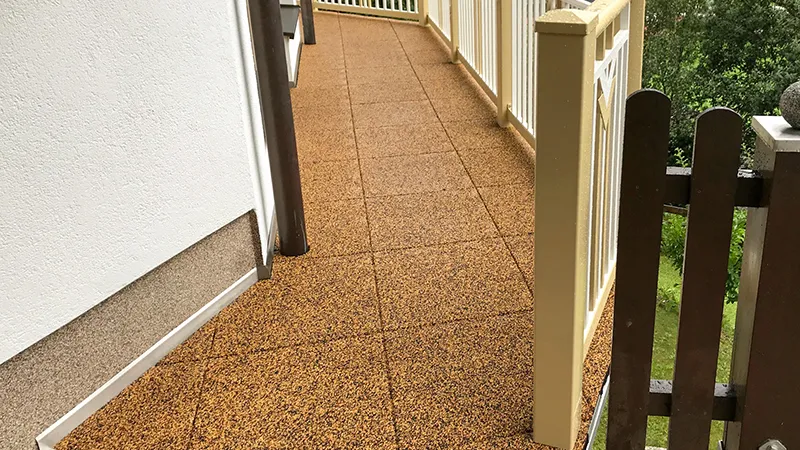 Podlouhlý balkon je pokryt balkonovými dlaždicemi WARCO v přirozeně působící béžovo-hnědé barvě. Po obvodu ho lemuje klasické, světlé zábradlí. Elastický povrch přesně přiléhá ke stěnám a sloupkům a chrání podkladovou konstrukci; gumová podlaha je příjemně pružná, protiskluzová a tím bezpečná i pohodlná pro děti, seniory a domácí mazlíčky.
