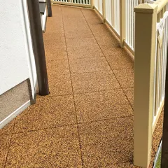 Podlouhlý balkon je pokryt balkonovými dlaždicemi WARCO v přirozeně působící béžovo-hnědé barvě. Po obvodu ho lemuje klasické, světlé zábradlí. Elastický povrch přesně přiléhá ke stěnám a sloupkům a chrání podkladovou konstrukci; gumová podlaha je příjemně pružná, protiskluzová a tím bezpečná i pohodlná pro děti, seniory a domácí mazlíčky.