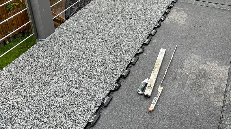 Na balkóně budou instalovány balkonové dlaždice WARCO z gumového granulátu. Skryté puzzle okraje zajišťují snadnou a přesnou pokládku. Vedle dlaždic jsou připraveny stahovací lať a metr k ověření rovnosti povrchu, který je utěsněn asfaltovou lepenkou.