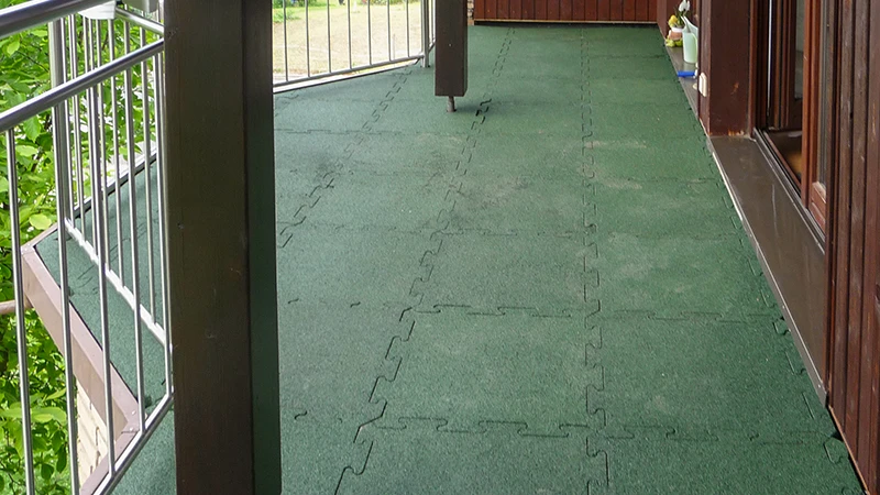 Na fotografii je dlouhý balkon pokrytý zelenými WARCO dlaždicemi v puzzle provedení. Protiskluzový a pružný povrch zaručuje bezpečí a pohodlí i za vlhka. Díky přesnému systému spojování vzniká odolné a snadno udržovatelné řešení, které chrání podklad. Výsledkem je příjemný venkovní prostor, ideální k relaxaci a vychutnání čerstvého vzduchu.
