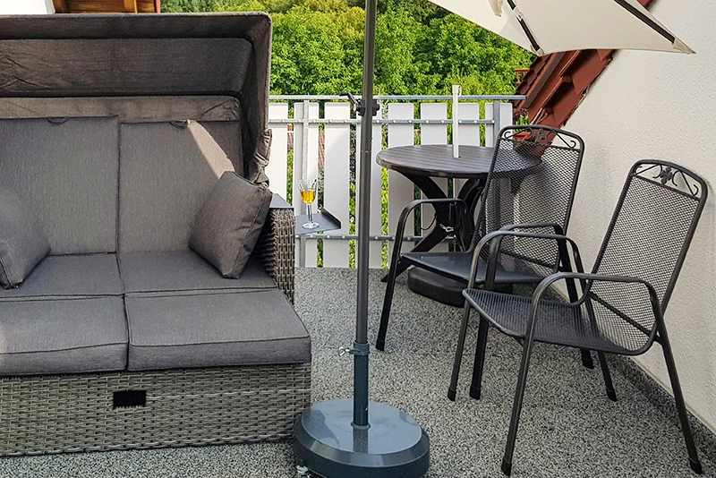 Na fotografii je balkon pokrytý šedými dlaždicemi WARCO. Pohodlné plážové křeslo a malý stolek se dvěma kovovými židlemi tvoří ideální místo k odpočinku a vychutnání výhledu. Elastický, protiskluzový povrch chrání před vlhkostí a tlumí kroky. Zároveň je snadno udržovatelný a odolný vůči povětrnostním vlivům, takže venkovní prostor zůstává příjemný po dlouhou dobu.