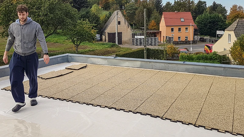 Na fotografii je vidět osoba, která pokládá béžové puzzle dlaždice WARCO na prostorný balkon. Díky přesnému systému spojování jde instalace rychle a snadno. Protiskluzový a povětrnostně odolný povrch chrání podklad a vytváří pochozí zónu pro různé využití. Výsledkem je bezpečný, pohodlný a snadno udržovatelný venkovní prostor, ideální k odpočinku. Navíc izolační účinek pomáhá snižovat hluk kroků.