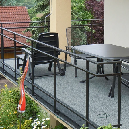 Na fotografii je prostorný balkon, pokrytý šedými dlaždicemi WARCO. Černé kovové zábradlí ohraničuje prostor a zajišťuje bezpečnost. Velký stůl a židle lákají k posezení. Elastická, protiskluzová podlaha je odolná vůči povětrnostním vlivům a snadno se udržuje, což zaručuje dlouhodobou ochranu a pohodlné místo k odpočinku na čerstvém vzduchu.