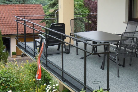 Na fotografii je prostorný balkon, pokrytý šedými dlaždicemi WARCO. Černé kovové zábradlí ohraničuje prostor a zajišťuje bezpečnost. Velký stůl a židle lákají k posezení. Elastická, protiskluzová podlaha je odolná vůči povětrnostním vlivům a snadno se udržuje, což zaručuje dlouhodobou ochranu a pohodlné místo k odpočinku na čerstvém vzduchu.