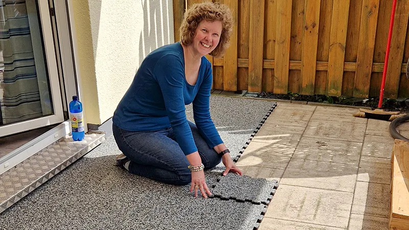 Domácí kutilka zde pokládá na svou terasu podlahu WARCO z PU pojeného gumového granulátu. Díky pevnému puzzle spojení lze elastické dlaždice rychle přizpůsobit a položit bez dalších nástrojů. Tento odolný a protiskluzový povrch chrání proti povětrnostním vlivům, zachovává si atraktivní vzhled a poskytuje vysoký komfort při chůzi.