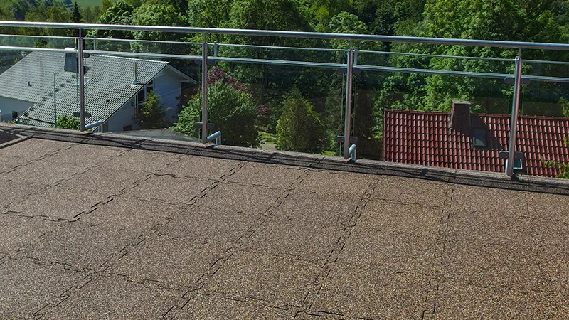 Na fotografii je vidět rozlehlý balkon s průhledným zábradlím, odkud se naskýtá výhled na okolní střechy a bujnou zeleň. Podlaha je pokryta puzzle dlaždicemi WARCO z gumového granulátu v teplém hnědém odstínu. Díky vzájemně se proplétajícím okrajům vzniká jednolitý povrch, který je protiskluzový a odolný vůči povětrnostním vlivům—ideální pro venkovní prostory. Navíc dlaždice tlumí nárazy a poskytují dobrou zvukovou izolaci. Snadná údržba z nich dělá odolné a pohodlné řešení pro balkony a terasy.